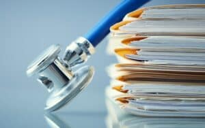 A stethoscope rests next to a stack of healthcare documentation and folders on a reflective surface.
