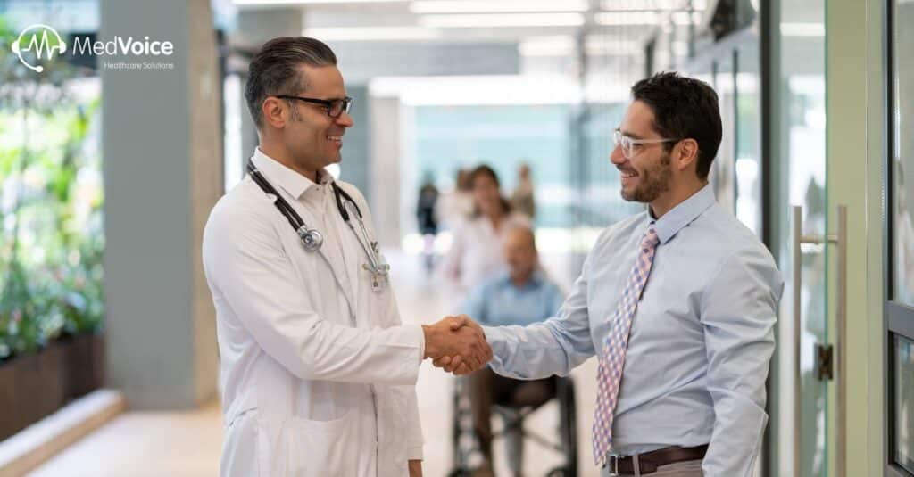 A doctor and a man in business attire shake hands in a hospital corridor, reflecting successful healthcare credentialing, with other people and a person in a wheelchair visible in the background.