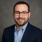 A man with glasses and a beard, wearing a blue shirt and dark blazer, smiles confidently like he's ready to launch the next big RCM initiative, set against a sleek gray background perfect for a professional landing page.