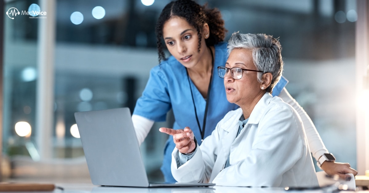 Two healthcare professionals, one in blue scrubs and one in a white coat, review Healthcare KPIs on a laptop screen together in a clinical setting.