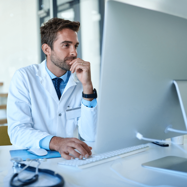 A doctor in a white coat sits at a desk, looking thoughtfully at a large computer monitor while using a keyboard and mouse—reviewing Healthcare KPIs related to Revenue Cycle Management. A stethoscope and documents are on the desk.