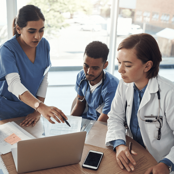 Three healthcare professionals review documents and a laptop at a table in a bright office, discussing Healthcare KPIs. One points at the screen while the others listen and take notes.