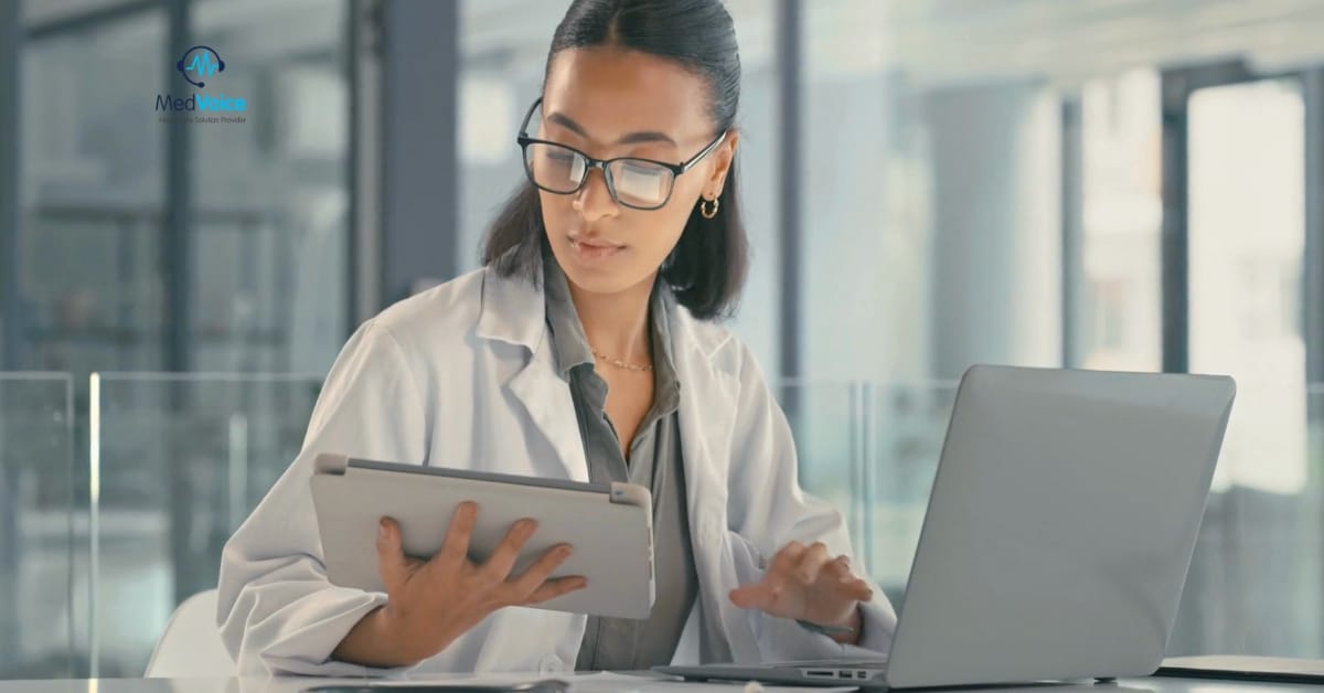 A person in a white lab coat uses a tablet and a laptop at a desk in a modern office, with the MedVoice logo visible in the background, highlighting advanced medical billing systems in use.