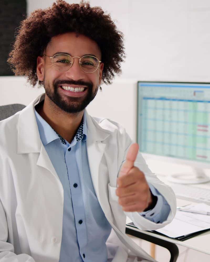 A smiling person in a white lab coat and glasses gives a thumbs-up while sitting at a desk, with a computer displaying a Medical Billing Review spreadsheet in the background.