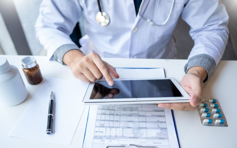 A doctor in a white coat uses a tablet at a desk with medical documents, a pen, a pill bottle, and a blister pack of pills—streamlining healthcare documentation.