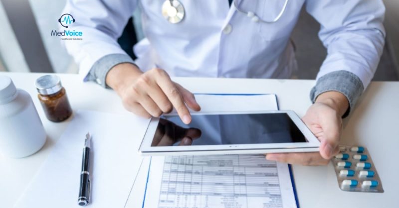 A doctor in a white coat uses a tablet at a desk with medical documents, a pen, pill bottles, and a blister pack of medication.