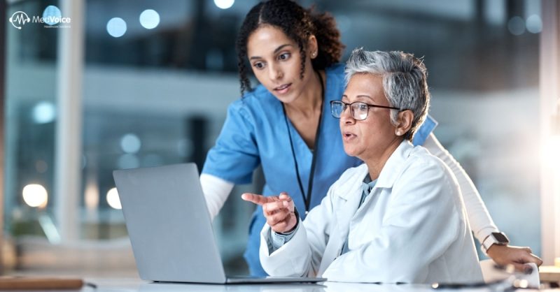 Two healthcare professionals, one in blue scrubs and one in a white coat, review Healthcare KPIs on a laptop screen together in a clinical setting.