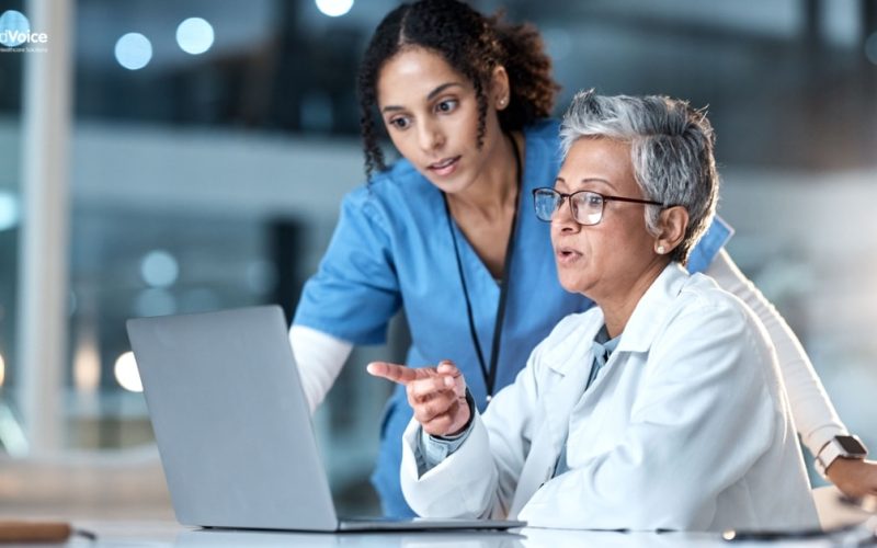 Two healthcare professionals, one in blue scrubs and one in a white coat, review Healthcare KPIs on a laptop screen together in a clinical setting.