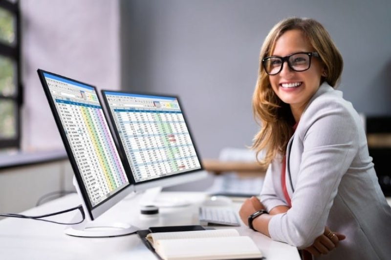 A woman wearing glasses sits at a desk with two computer monitors displaying spreadsheets, smiling at the camera. A notebook and pen are on the desk, reflecting her role in US healthcare and AI in revenue cycle management.