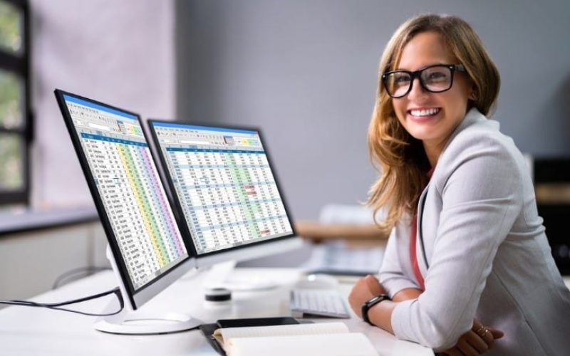 A woman wearing glasses sits at a desk with two computer monitors displaying spreadsheets, smiling at the camera. A notebook and pen are on the desk, reflecting her role in US healthcare and AI in revenue cycle management.