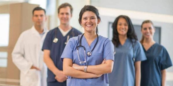 Five healthcare professionals in scrubs and lab coats stand together indoors, with one woman in blue scrubs smiling at the front, representing the MedVoice team’s dedication to patient care.