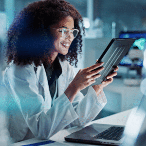A scientist wearing a lab coat and glasses sits at a desk, smiling while using a tablet; a laptop is open in front of her in a modern laboratory setting, where she's focused on accurate coding and reviewing E/M codes.