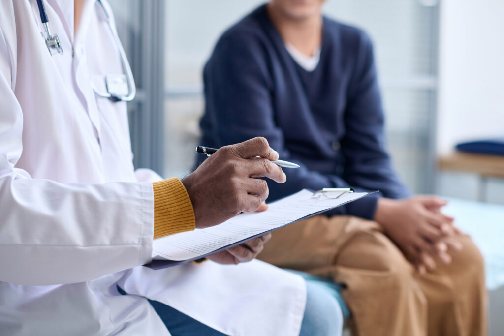 A doctor holds a clipboard and pen, reviewing medical records while speaking with a patient seated nearby in an examination room.