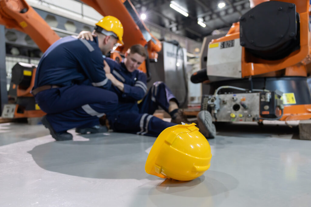 A yellow hard hat lies on the floor while two workers in safety gear attend to an injured colleague in an industrial setting with robotic machinery, highlighting the importance of medical review protocols.
