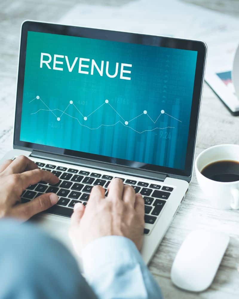 Person working on a laptop displaying "REVENUE" and a line graph—perfect for illustrating RCM review or revenue cycle management; coffee and a mouse are on the desk.