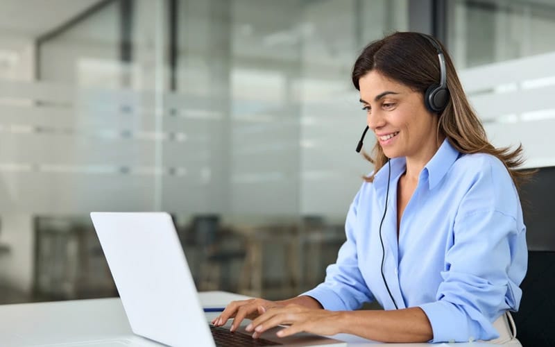A woman wearing a MedVoice headset sits at a desk, smiling, and types on a laptop in a modern office setting.