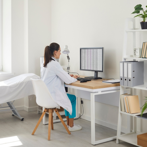 A doctor in a white coat sits at a desk in a medical office, working on a computer displaying a spreadsheet to review the medical billing benefits for independent practices.
