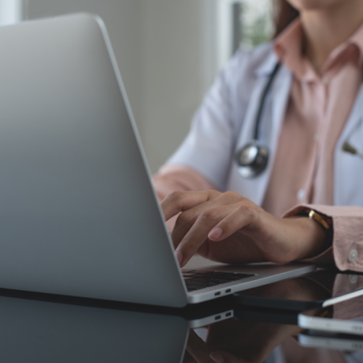 A person wearing a white coat and stethoscope types on a laptop at a desk, suggesting they are a healthcare professional working, possibly exploring the benefits of outsourcing medical billing for independent practices.