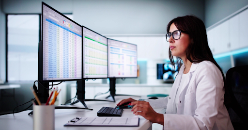 A woman in a white lab coat works at a desk with three large computer monitors displaying spreadsheets and data charts, highlighting the medical billing benefits for independent practices.