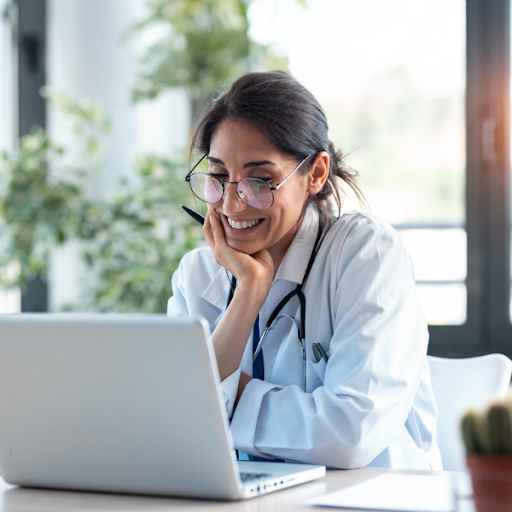 A doctor wearing a white coat and stethoscope smiles while looking at a laptop, sitting at a desk in a bright office.