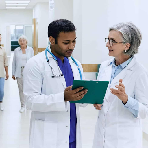 Two doctors in white coats discuss patient notes in a hospital corridor, while two people walk in the background.