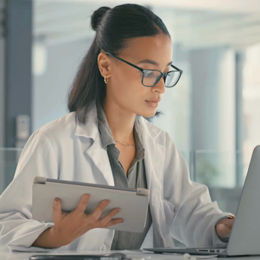 A woman wearing a white lab coat and glasses works at a desk, holding a tablet in one hand and typing on a laptop with the other.