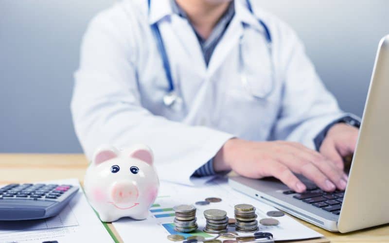 A doctor focused on RCM sits at a desk, working on a laptop surrounded by charts, a piggy bank, coins, and a calculator.